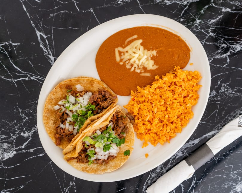 Two tacos with rice and refried beans on a white plate, on a black marble table.