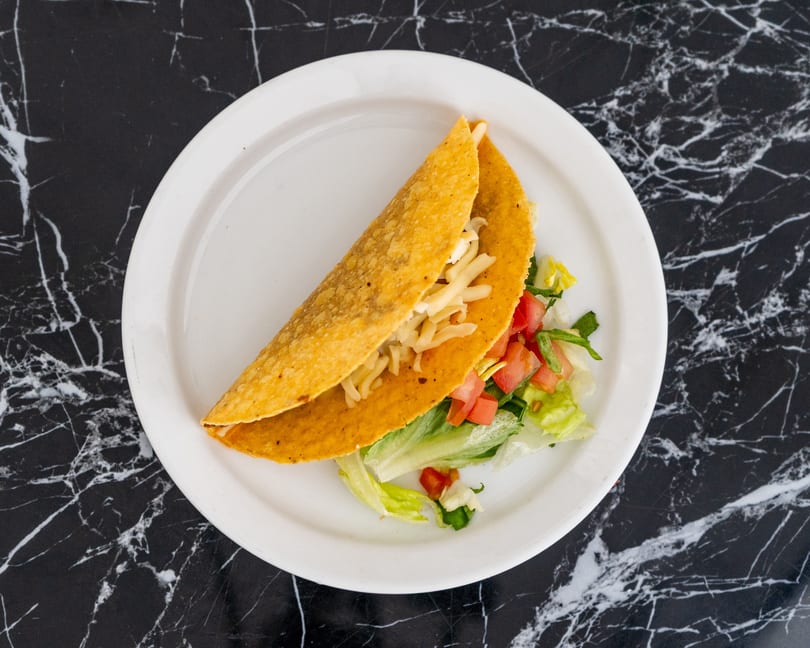 A single hard-shell taco filled with meat, shredded cheese, lettuce, and diced tomatoes on a white plate.